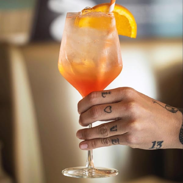 A tattooed hand holding a glass of Pecoraro Latteria's Aperol Spritz with an orange slice.
