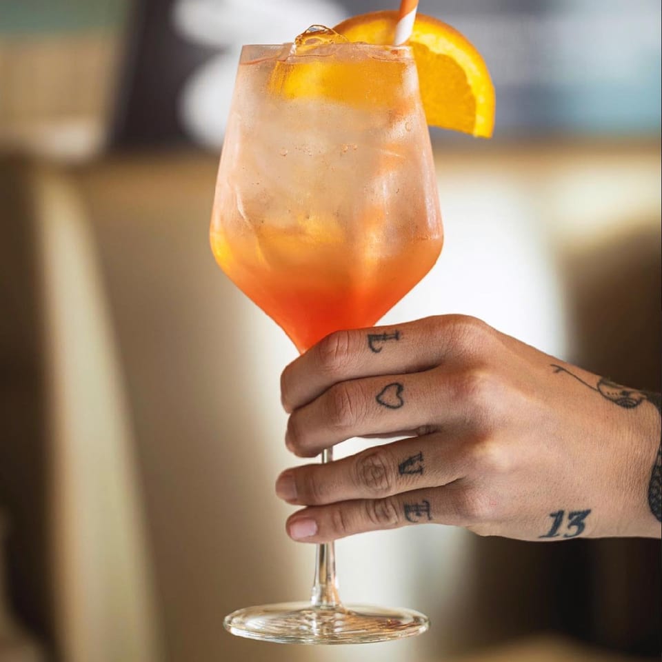 A tattooed hand holding a glass of Pecoraro Latteria's Aperol Spritz with an orange slice.