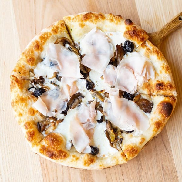 Italian pizza with white sauce, mushrooms, black olives, and prosciutto on a wooden paddle.