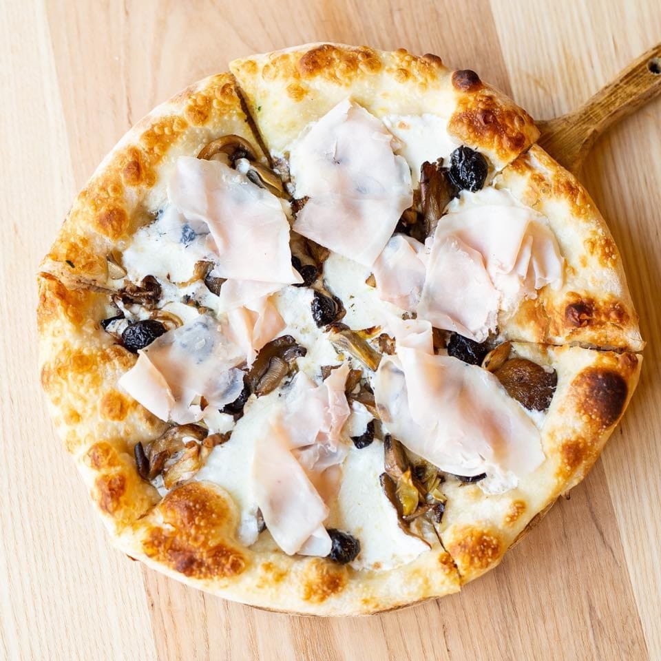 Italian pizza with white sauce, mushrooms, black olives, and prosciutto on a wooden paddle.