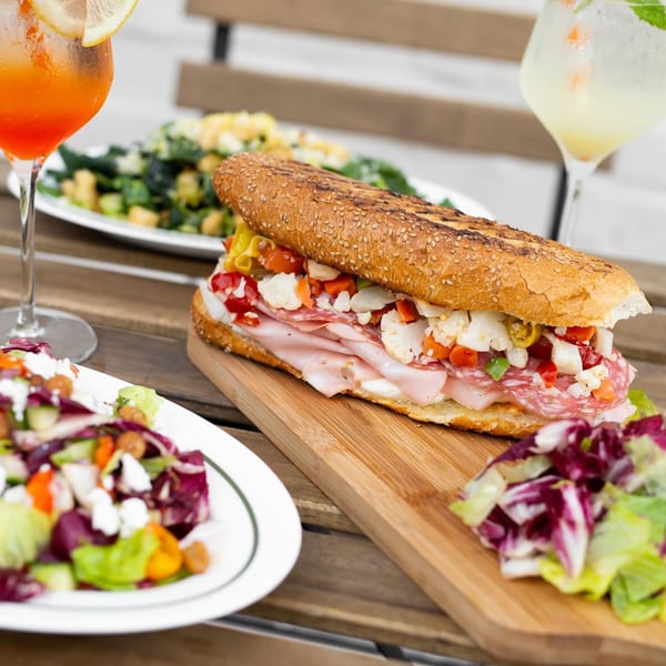 Italian sandwich with meats, cheese, and pickled vegetables, served with salads and cocktails on a wooden table.