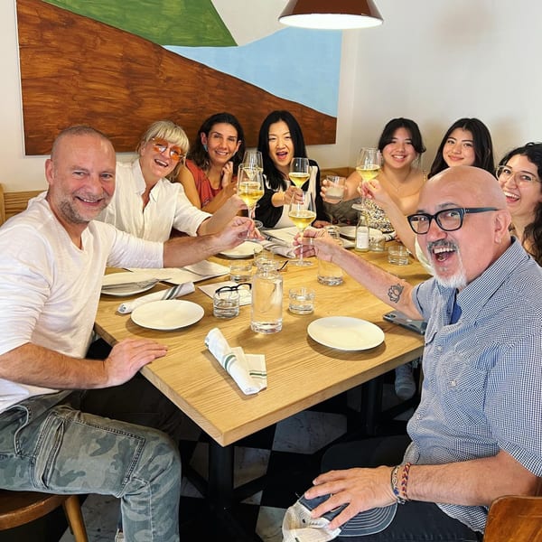 Eight people toasting with white wine glasses around a wooden table in an Italian restaurant.