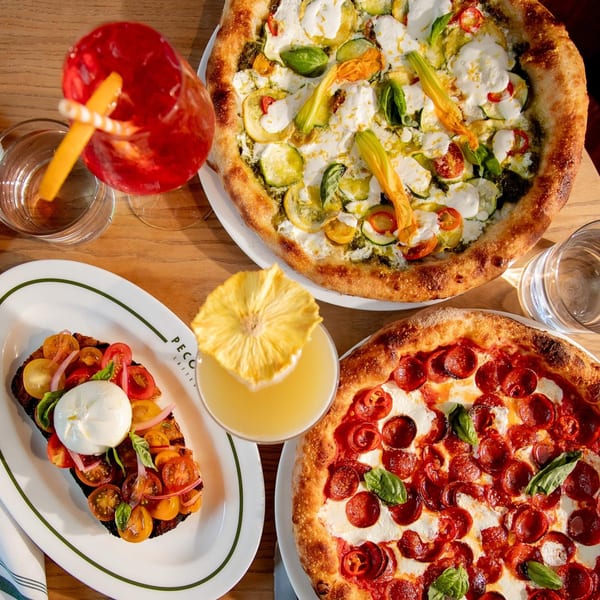 Two Italian pizzas, bruschetta with burrata, and two cocktails on a wooden table.