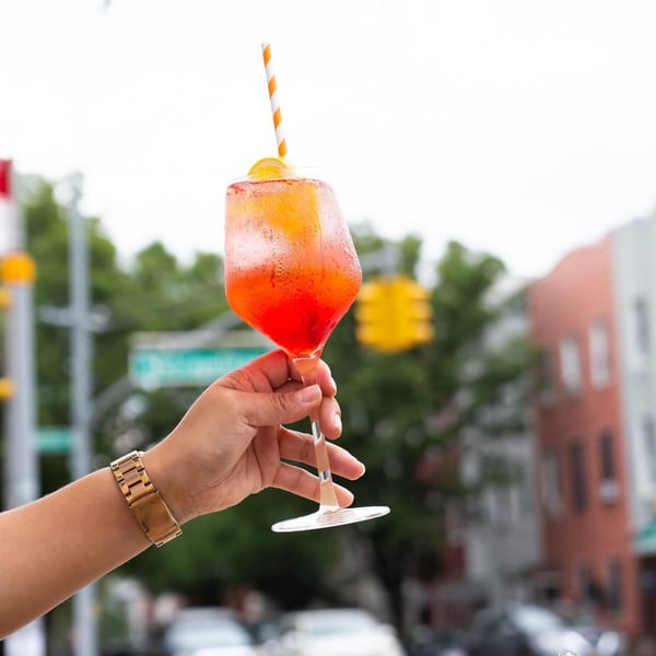 A hand holding up a vibrant orange and red Aperol Spritz with a straw and orange slice.