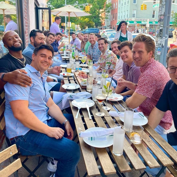 A group of men smiling and dining al fresco at a long wooden table at Pecoraro Latteria.